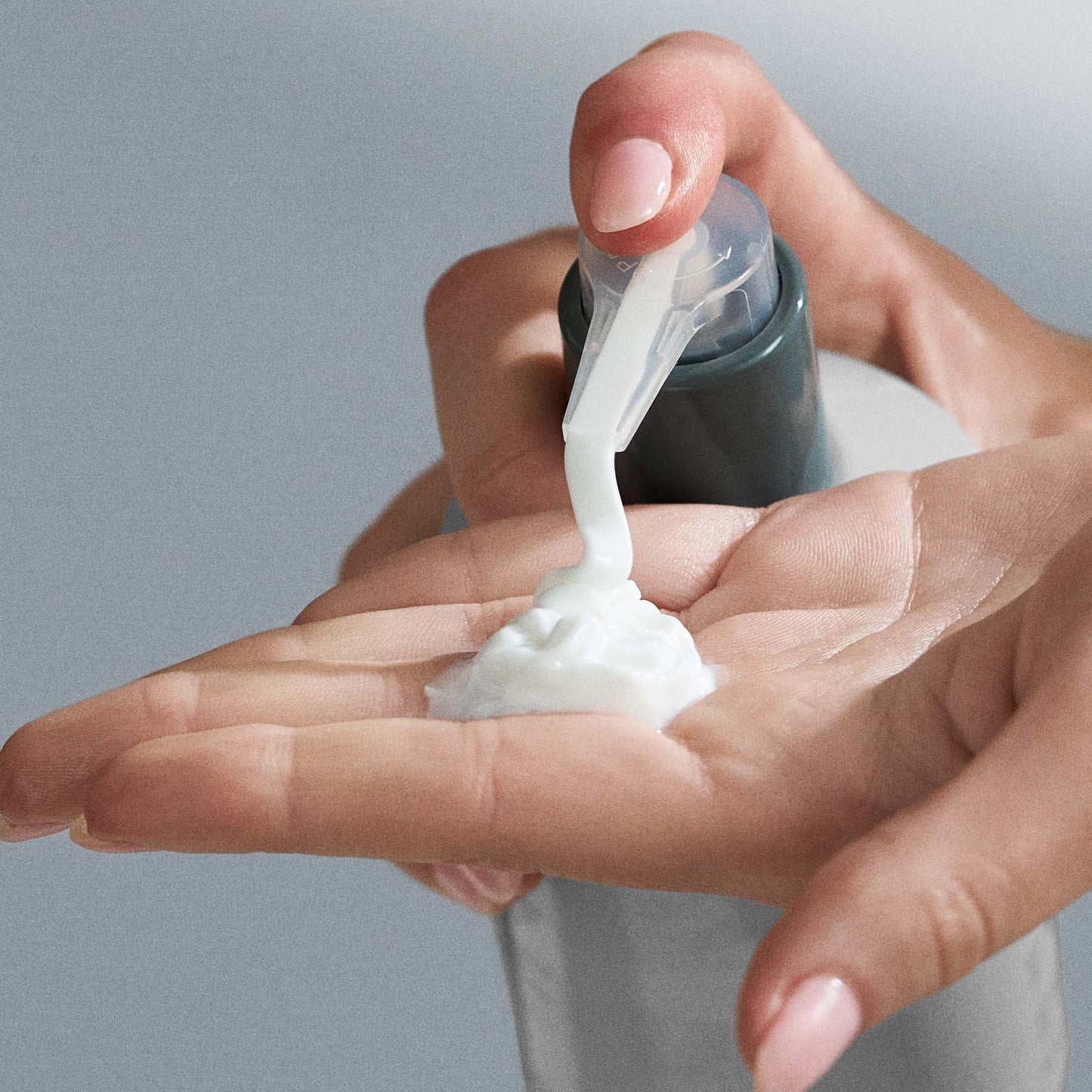 Hand applying a white cream from a bottle onto another hand against a neutral background 